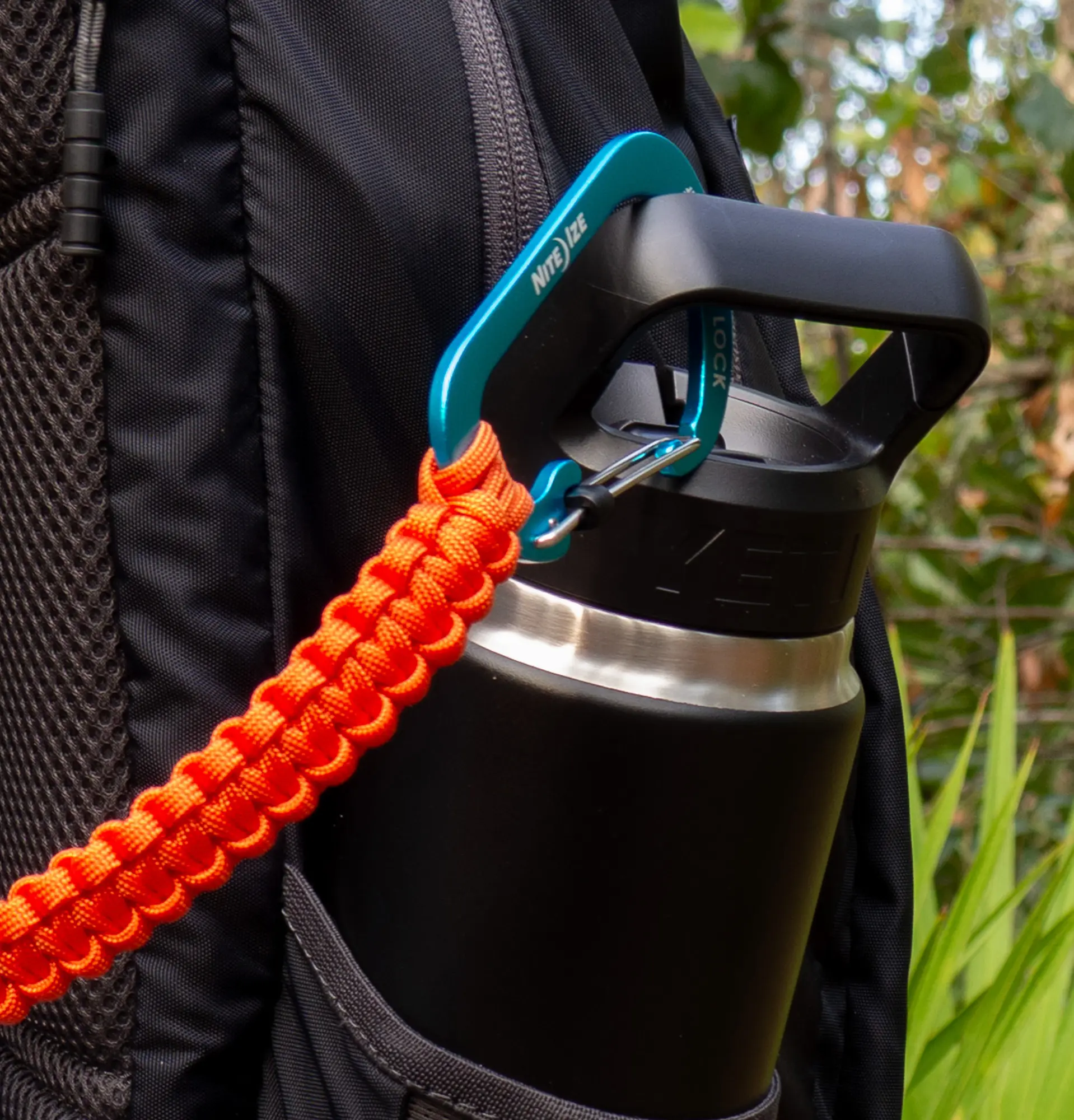 Close-up of an orange THIRSTLOCK™ ANCHOR™ bottle leash with blue carabiner attached to a water bottle in a backpack pocket.
